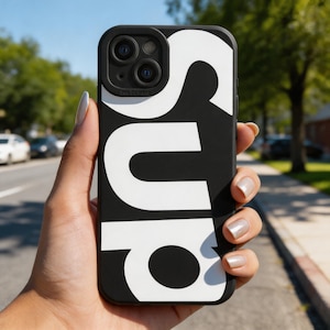May include: Black phone case with the word "SUP" in large white letters. The case is held in a hand against a background of a street and trees.