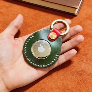 May include: A green leather keychain holder with white stitching, a red accent, and a gold-colored ring. The holder encases a silver Apple AirTag with the Apple logo. The keychain is designed to hold a tracking device.