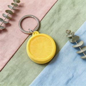 May include: A yellow, circular keychain with a silver key ring. The keychain has a textured surface and a small loop at the top. The background is a mix of pink, green, and blue fabric with sprigs of greenery.