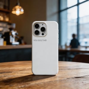 May include: A white phone case with a black camera module and the text "YOU GOS THIS" printed on the back. The phone case is sitting on a wooden table in a cafe setting.