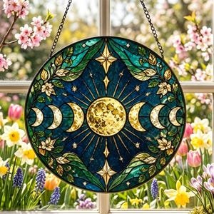 May include: A circular stained glass suncatcher featuring a central yellow moon surrounded by a lunar cycle of crescent and gibbous phases. The design includes deep blue glass, green foliage, and white flowers, all framed by a black metal chain.