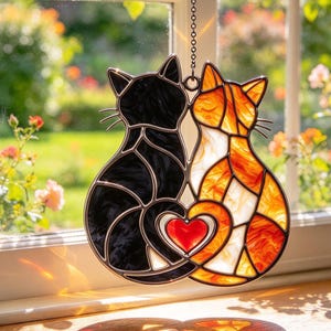 May include: A stained glass suncatcher featuring two cats sitting side by side, one in black and one in orange and white, with their tails forming a heart shape around a central red glass heart. The piece hangs from a metal chain.