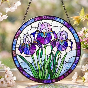 May include: A circular stained glass suncatcher featuring three purple and white iris flowers with green stems and leaves. The design is enclosed in a dark metal frame and suspended by a black chain, showcasing vibrant glass textures and colours.