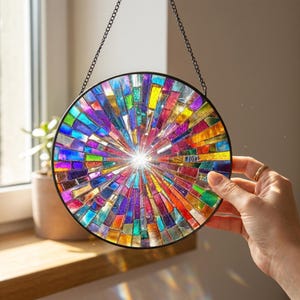 May include: A circular suncatcher featuring a vibrant mosaic of multicolored glass shards arranged in a radial pattern. The glass pieces include shades of blue, purple, red, orange, yellow, and green, all encased in a thin black metal frame.