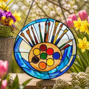 Könnte beinhalten: Ein runder Buntglas-Sonnenfänger mit einer zentralen Künstlerpalette, die acht farbige Farbkreise in Rot, Gelb, Blau, Grün und Orange zeigt. Um die Palette herum sind verschiedene Pinsel mit Holzgriffen und Borsten angeordnet.