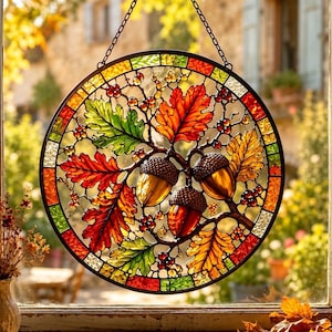 Op de afbeelding: Een ronde glas-in-lood suncatcher met een gedetailleerd herfstmotief van drie bruine eikels, levendige rode, oranje en groene eikenbladeren en kleine rode bessen. Het stuk is gevat in zwart metaal en hangt aan een dunne metalen ketting.