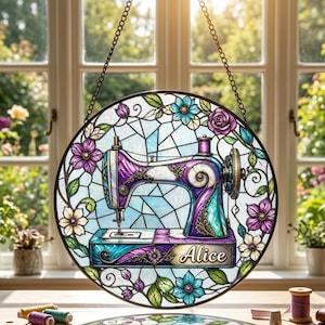 May include: A circular stained glass suncatcher featuring a purple and teal sewing machine with the name Alice printed on the base. The design is surrounded by a border of colorful flowers and green leaves, suspended by a metal chain.