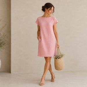 May include: A knee-length, short-sleeved dress in a solid light pink color. The garment features a simple round neckline, a straight-cut silhouette, and two side pockets. The fabric has a visible, slightly textured weave throughout the entire piece.