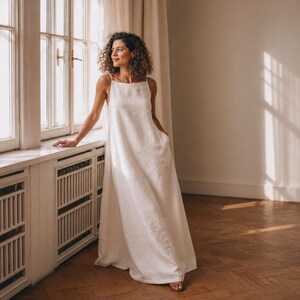 Könnte beinhalten: Ein bodenlanges, ärmelloses weißes Kleid mit quadratischem Ausschnitt und dünnen Trägern. Das Kleidungsstück hat eine lockere, fließende Silhouette, die bis zum Boden reicht, und ist aus einem strukturierten Stoff mit sichtbaren Seitentaschen gefertigt.