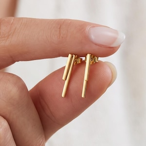 Könnte beinhalten: Ein Paar goldfarbene Ohrstecker mit zwei parallelen, vertikalen Stäben unterschiedlicher Länge. Die Ohrringe haben eine polierte metallische Oberfläche und werden zwischen den Fingern gehalten, was das schlanke, lineare Design unterstreicht.