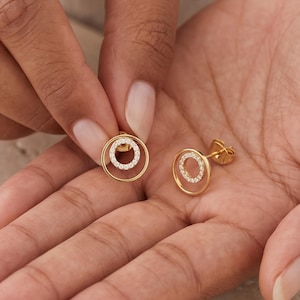 Könnte beinhalten: Ein Paar goldene Ohrstecker mit kreisförmigem Design. Jeder Ohrring besteht aus einem größeren äußeren Goldring und einem kleineren inneren Ring, der mit kleinen klaren Steinen besetzt ist. Die Ohrringe haben ein poliertes Finish und einen Stecker.
