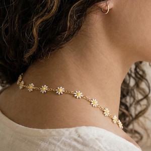 Könnte beinhalten: Eine goldfarbene Halskette mit Gänseblümchen-Kette mit weißen und gelben Blumendetails. Die Halskette wird um den Hals getragen. Die Person trägt ein cremefarbenes Hemd.