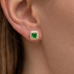 Puede incluir: Pendiente de botón de esmeralda de corte cuadrado con un halo de pequeños diamantes transparentes. La esmeralda es de un verde vibrante y los diamantes brillan sobre el engaste de oro. El pendiente se muestra en una oreja.