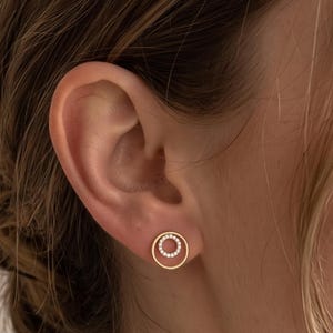 Puede incluir: Un pendiente de botón en oro con un pequeño marco circular y un círculo interior adornado con diminutas piedras transparentes. El diseño consiste en un anillo exterior de oro pulido que rodea un círculo interior más pequeño con incrustaciones.