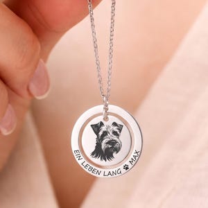 Może przedstawiać: Srebrzysty, okrągły naszyjnik z zawieszką z wygrawerowanym wizerunkiem głowy psa w centrum. Zewnętrzny pierścień zawiera napis "EIN LEBEN LANG MAX" oraz mały symbol odcisku łapy. Zawieszka jest umieszczona na łańcuszku.