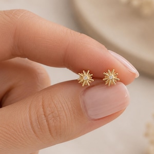Könnte beinhalten: Ein Paar goldene Ohrstecker in Form von Sonnenstrahlen, jeweils mit einem kleinen, klaren, rund geschliffenen Stein in der Mitte. Die Ohrringe haben ein strahlendes, sternförmiges Design mit mehreren dünnen, spitzen Strahlen.