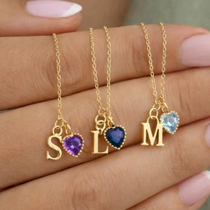 May include: Gold necklaces featuring initial letter pendants and heart-shaped gemstone charms. The letters are S, L, and M. Gemstones are purple, dark blue, and light blue. The necklaces are shown against a neutral backdrop.