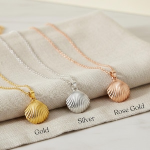 Könnte beinhalten: Drei Muschel-Anhänger-Halsketten in Gold, Silber und Roségold. Jeder Anhänger hat ein geripptes Design und hängt an einer zarten Kette. Die Halsketten werden auf einem neutralfarbenen Stoff präsentiert.
