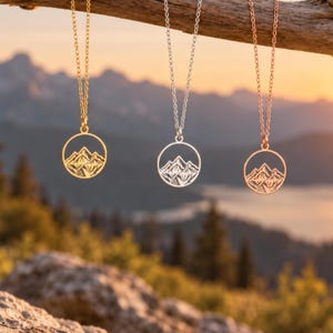 Puede incluir: Tres collares con colgantes circulares que presentan un diseño recortado de una cordillera. Un colgante es de tono dorado, otro de tono plateado y otro de tono oro rosa. Cada colgante cuelga de una fina cadena de metal a juego.