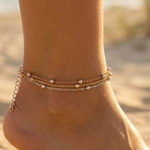 May include: A multi-strand gold-toned anklet featuring three delicate chains adorned with small, evenly spaced metallic beads. The jewelry piece includes a thin extender chain with a small clasp, designed to wrap around the ankle securely.