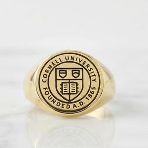 May include: A gold-toned signet ring featuring a circular face engraved with the Cornell University seal, including the text Cornell University and Founded A.D. 1865 surrounding a central shield emblem with an open book and crest details.