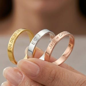 May include: Three thin metal rings in gold, silver, and rose gold tones. Each ring features custom engraving on the outer surface. The gold ring displays the date 06.06.2026, the silver ring shows the letters E and J with a heart, and the rose gold ring is engraved with the name Jack and a heart.