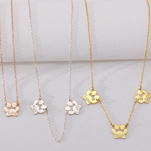 May include: A collection of delicate necklaces with paw print pendants. Each necklace features a chain and a paw print charm with a name engraved on it. The necklaces are in rose gold, silver, and gold tones.