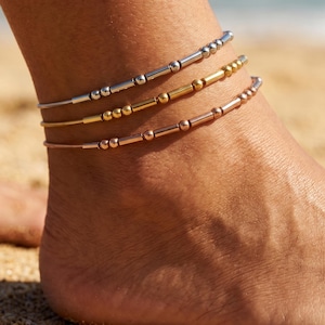 May include: Three delicate anklets worn together, each featuring a unique arrangement of small metallic beads and cylindrical spacers. The set includes one silver-toned, one gold-toned, and one rose gold-toned chain, all resting on a thin cord base.