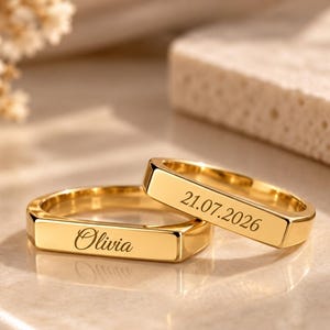 Könnte beinhalten: Zwei goldfarbene Metallringe mit flachen, rechteckigen Vorderseiten. Ein Ring ist mit dem Namen Olivia in Schreibschrift graviert, der andere Ring trägt das eingravierte Datum 21.07.2026 in einer serifenlosen Schrift.