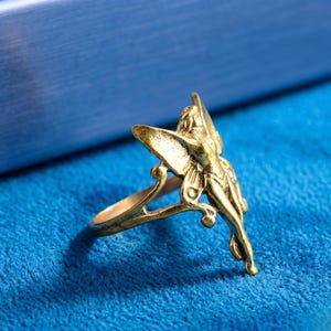 May include: A gold-coloured ring with a fairy design. The ring showcases a detailed depiction of a fairy with outstretched wings. The ring is set against a blue background, highlighting the intricate details of the jewellery.