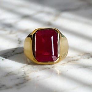 Puede incluir: Un anillo dorado con una gran gema rectangular de color rojo intenso. El anillo tiene una banda gruesa y un acabado pulido, que refleja la luz. La gema está engastada en un bisel. Un anillo llamativo.