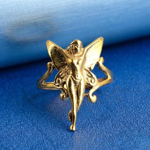 May include: A gold ring with a fairy design. The fairy has detailed wings and is the central focus of the ring. The ring is set against a blue background, showcasing the intricate details of the jewellery.