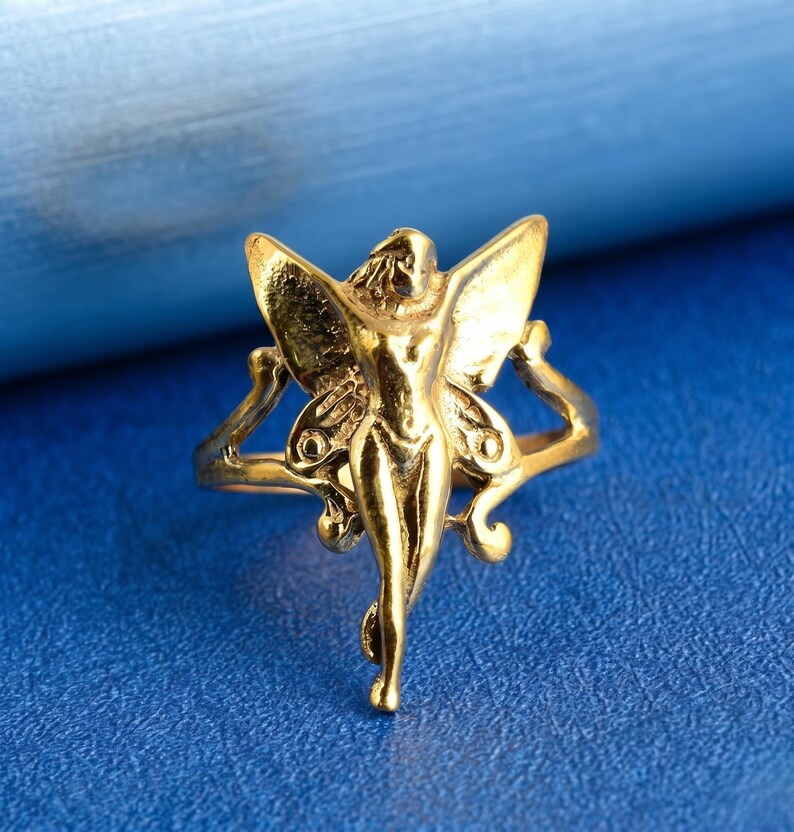 May include: A gold ring with a fairy design. The fairy has detailed wings and is the central focus of the ring. The ring is set against a blue background, showcasing the intricate details of the jewellery.