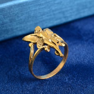 May include: A gold-coloured ring with a detailed fairy design. The ring's band curves gracefully, supporting the fairy figure with outstretched wings. The fairy is sculpted with intricate details, creating a whimsical and elegant piece of jewellery.