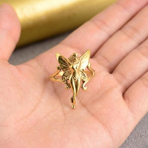May include: A gold-coloured ring with a fairy design. The fairy has outstretched wings and is in a standing pose. The ring is detailed with intricate patterns and is set against a neutral background.