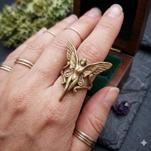 May include: A bronze-colored fairy ring with outstretched wings and a detailed figure. The ring is worn on a finger, showcasing its intricate design and metallic finish. The ring is a unique piece of jewelry.