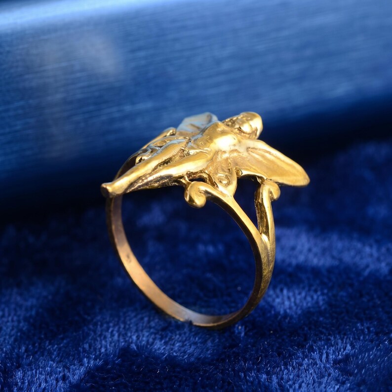 May include: A gold ring with a detailed sculpted figure with outstretched wings. The ring's design includes delicate curves and a small, light-coloured accent on the figure. The ring is set against a deep blue background.