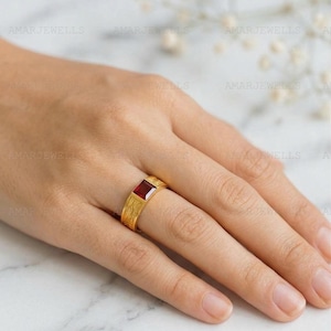 Può includere: Un anello color oro con una gemma rettangolare rosso scuro. L'anello ha una fascia strutturata e viene indossato su un dito. Lo sfondo è sfocato, con il marchio "AMARJEWELLS" visibile.