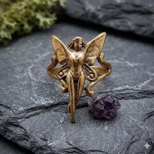 May include: A bronze-colored ring featuring a fairy design with outstretched wings. The fairy has a detailed face and body. A small, dark purple amethyst crystal sits nearby on a gray stone surface.