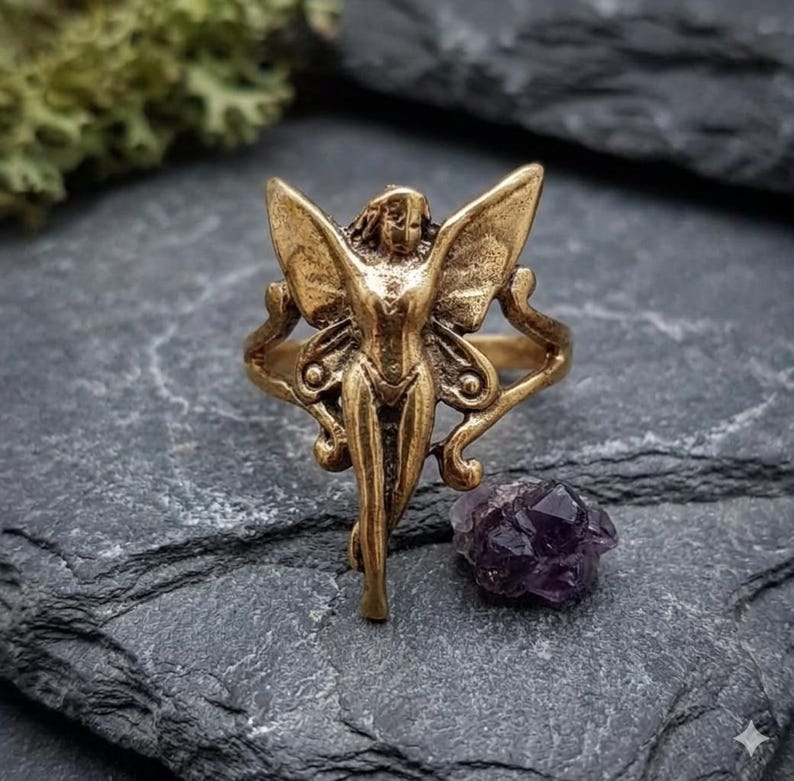 May include: A bronze-colored ring featuring a fairy design with outstretched wings. The fairy has a detailed face and body. A small, dark purple amethyst crystal sits nearby on a gray stone surface.