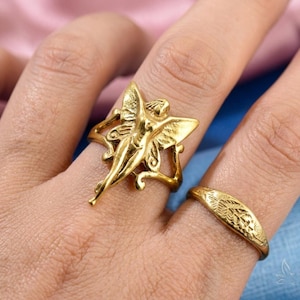May include: A gold ring with a fairy design and outstretched wings. The fairy ring is on a finger next to a second gold ring with a leaf pattern. The rings are on a light skin tone.