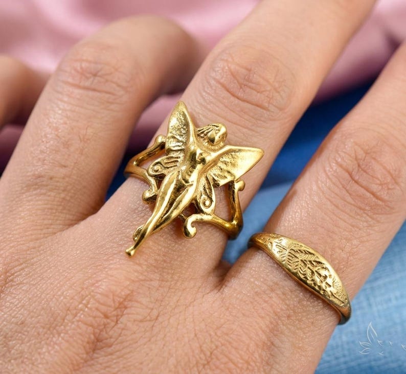 May include: A gold ring with a fairy design and outstretched wings. The fairy ring is on a finger next to a second gold ring with a leaf pattern. The rings are on a light skin tone.