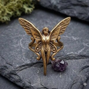 May include: A bronze-colored ring featuring a fairy design with outstretched wings. The ring has intricate details and is set against a dark, textured background. A small, purple crystal sits nearby.