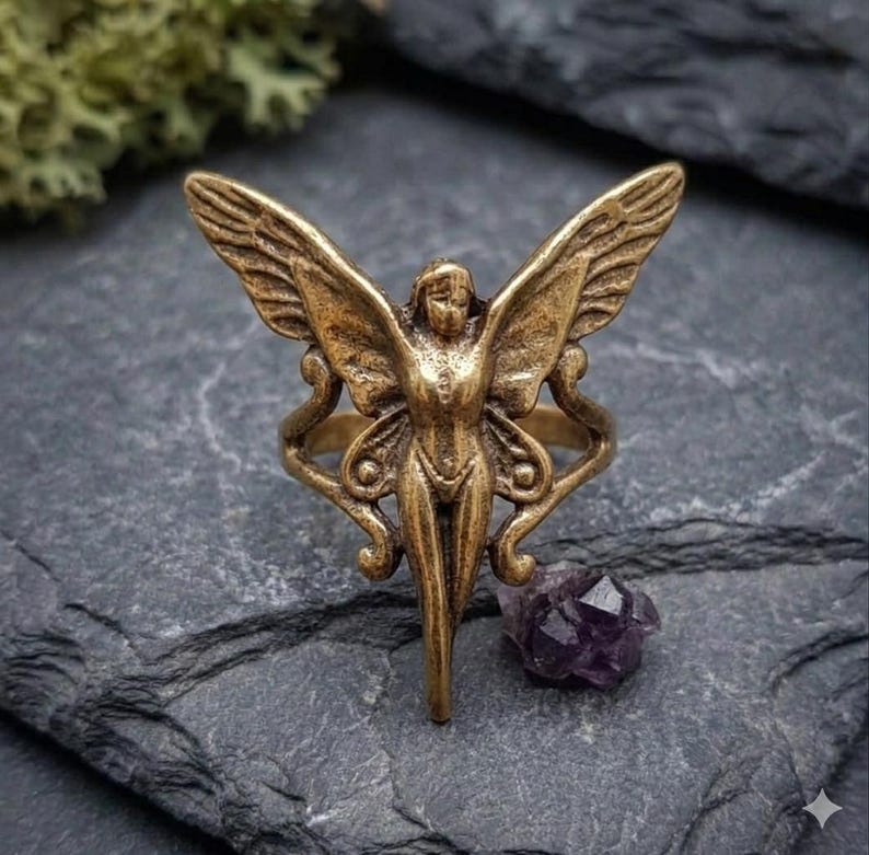 May include: A bronze-colored ring featuring a fairy design with outstretched wings. The ring has intricate details and is set against a dark, textured background. A small, purple crystal sits nearby.