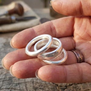 May include: Three silver rings with a smooth, rounded design are stacked in a hand. The rings have a polished finish, reflecting light. A textured copper-coloured ring is also visible on a finger. The background includes tools and a wooden surface.