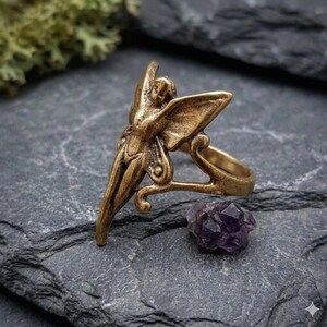 May include: A bronze-colored ring featuring a fairy design with outstretched wings. The ring has intricate details and is set against a dark gray stone surface. A small, purple amethyst crystal sits nearby, adding a touch of color.