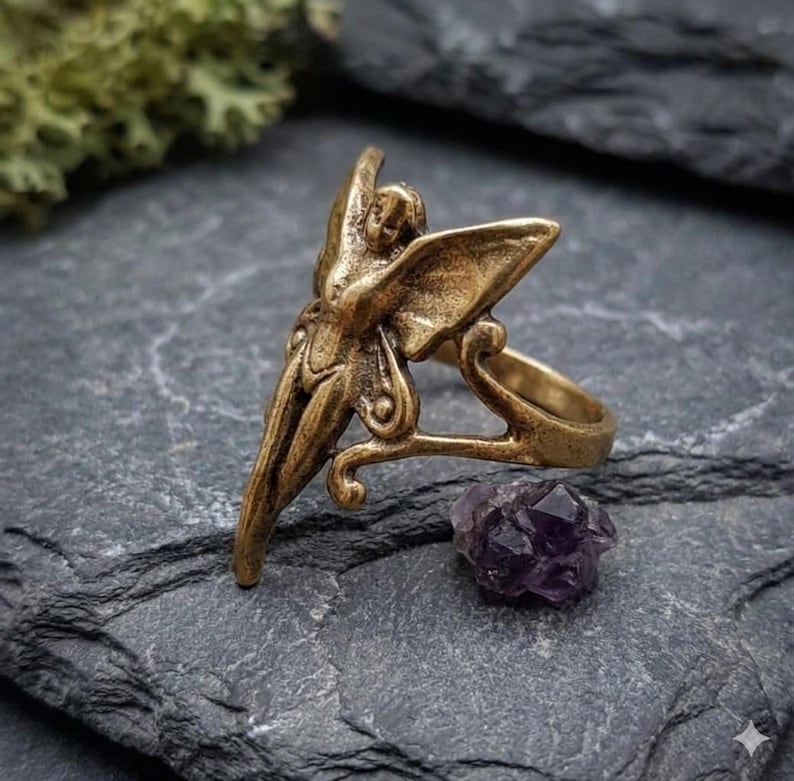 May include: A bronze-colored ring featuring a fairy design with outstretched wings. The ring has intricate details and is set against a dark gray stone surface. A small, purple amethyst crystal sits nearby, adding a touch of color.