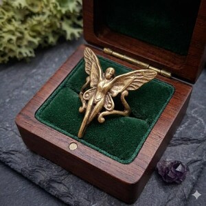 May include: A gold-colored ring in the shape of a winged figure, displayed in an open wooden box with a green velvet interior. The ring has intricate details and is set against a dark background. A small purple crystal is visible.