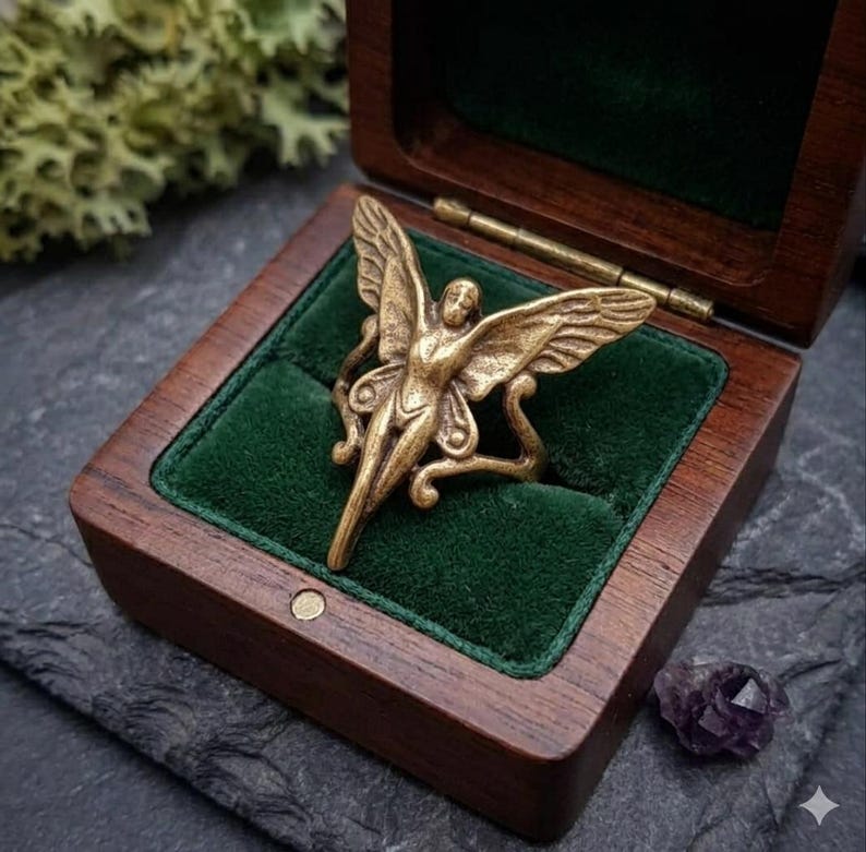 May include: A gold-colored ring in the shape of a winged figure, displayed in an open wooden box with a green velvet interior. The ring has intricate details and is set against a dark background. A small purple crystal is visible.