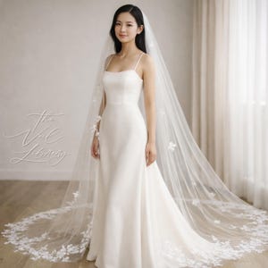 Puede incluir: Un vestido de novia blanco con una larga cola y un velo a juego. El velo está adornado con delicados apliques florales. El vestido tiene un diseño sencillo con tirantes finos. El texto "The Veil Library" es visible.